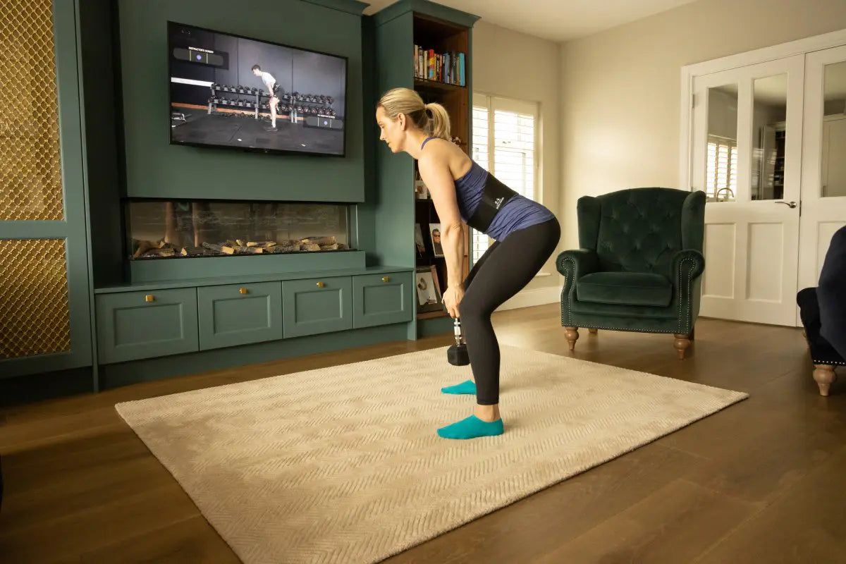 A woman hinging forward with a dumbbell in one hand, following a class on the television.