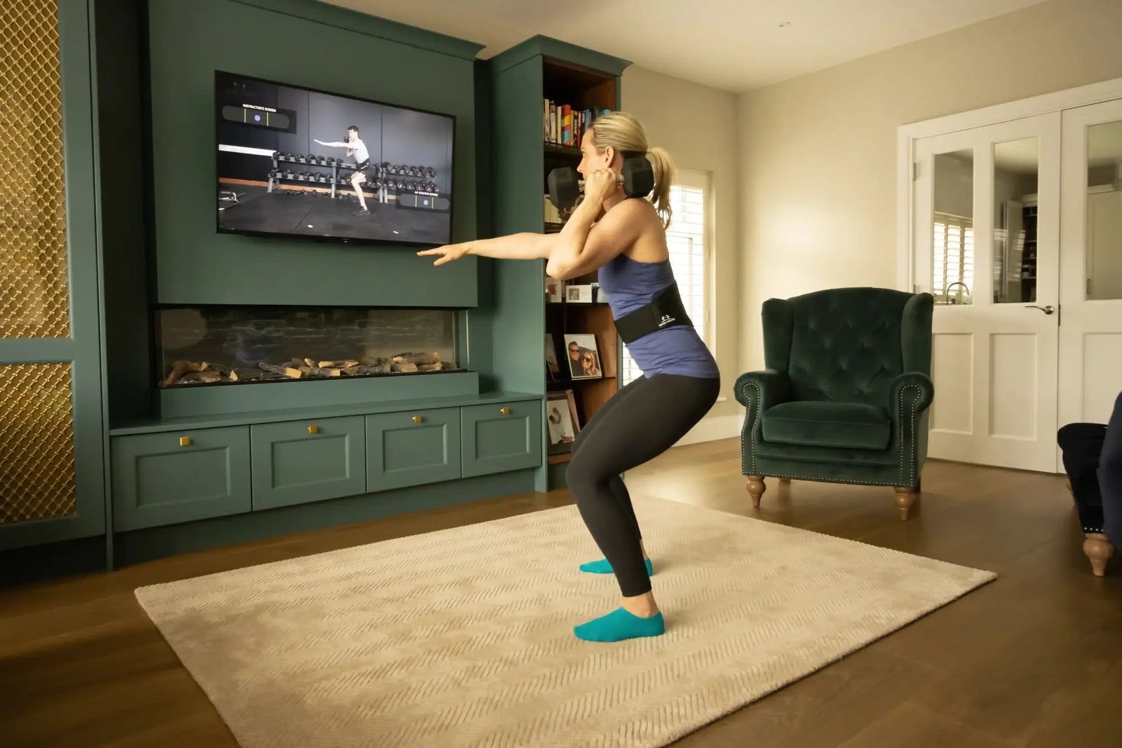 A woman wearing the belt in a squat with both arms reaching forward, following a class on the television.