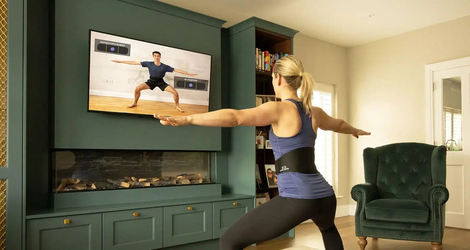 A woman with both arms stretched out to the sides, mirroring the instructor on the wall screen in front of her, the belt at her waist.