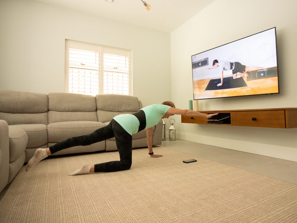 A woman on all fours on a rug, one arm and the opposite leg extended in a bird-dog, a class playing on the wall screen.
