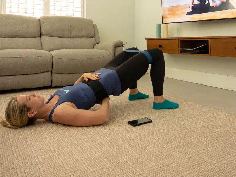 A woman on her back on a rug in a living room, hips lifted into a bridge, her phone on the floor beside her.