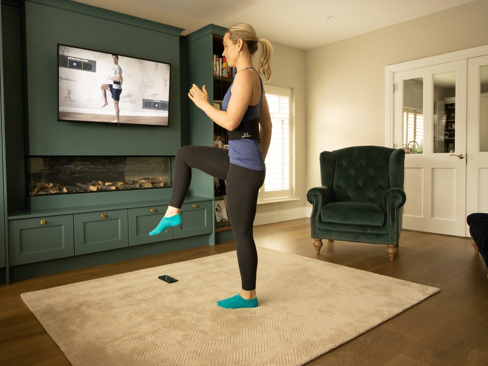 A woman standing on one leg with the other knee raised, following an instructor on the wall screen.