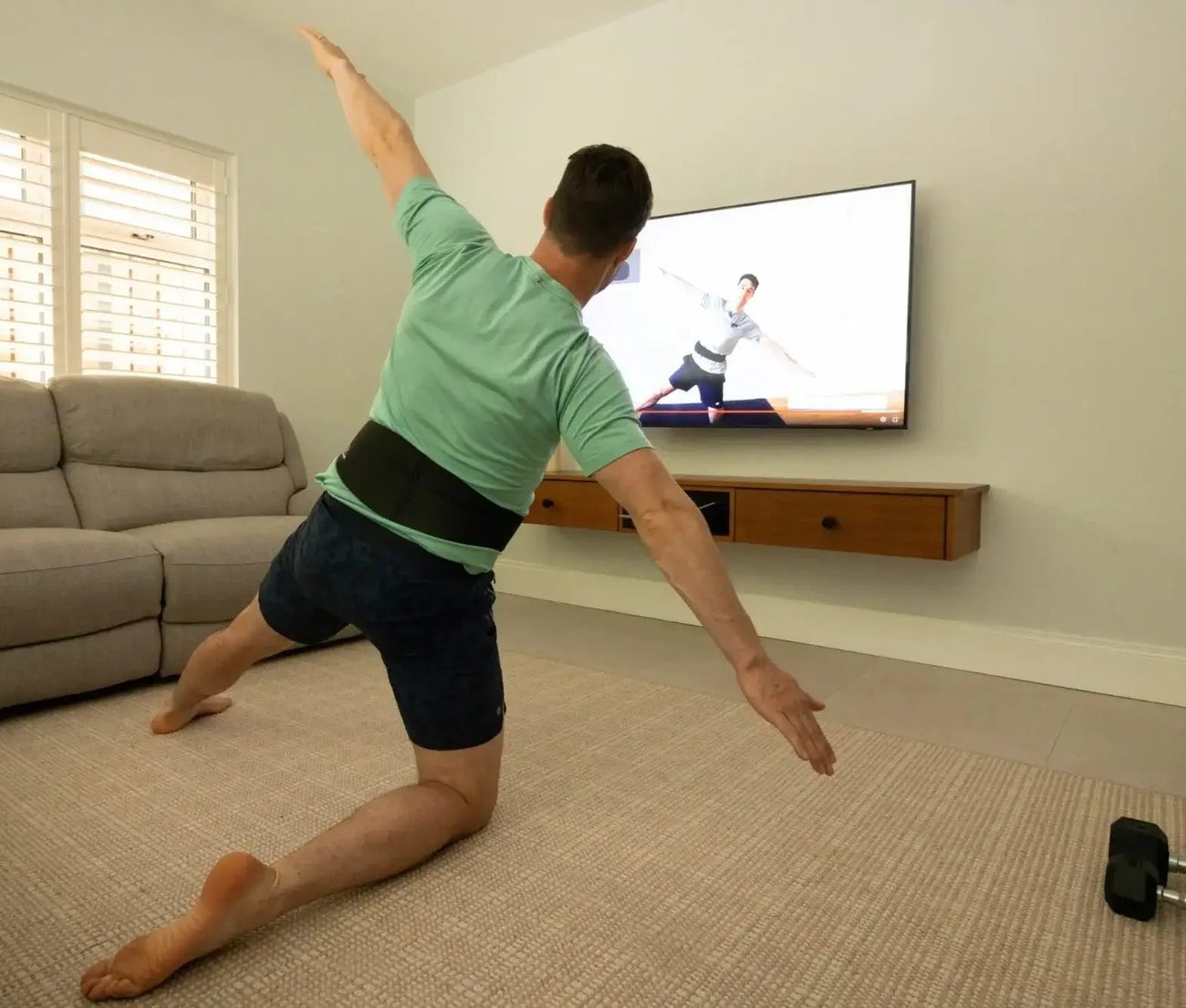 A man in a green t-shirt in a deep kneeling lunge with one arm raised overhead, the belt clearly visible at his waist, a class playing on the screen beside him.